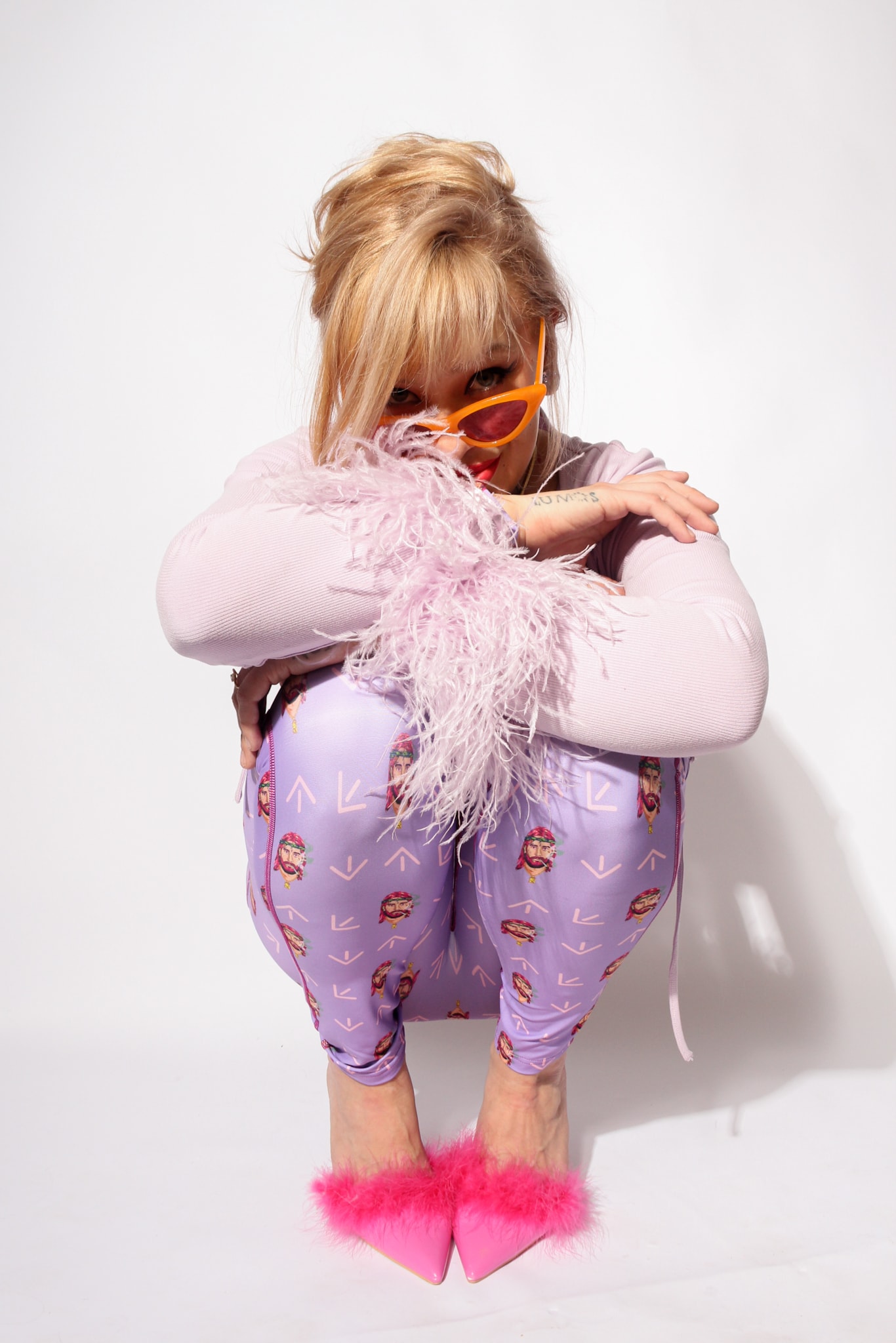 South North Southwest crouching in a lilac catsuit with her face hidden behind a feather boa